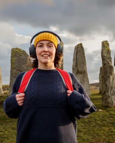 Female with a yellow hat, black overhead earphones, and a red backpack standing in front of ancient standing stones