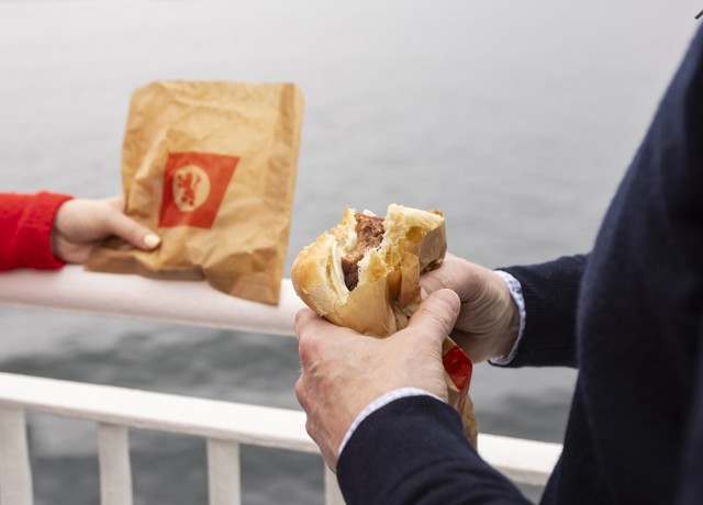 a girl in a red sweater holding a CalMac branded paper bag while a man in a navy jumper eats a slice sausage roll. 