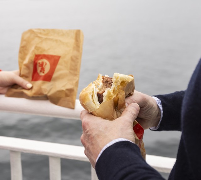 a girl in a red sweater holding a CalMac branded paper bag while a man in a navy jumper eats a slice sausage roll. 