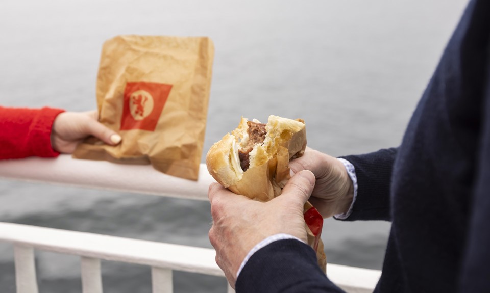 a girl in a red sweater holding a CalMac branded paper bag while a man in a navy jumper eats a slice sausage roll. 
