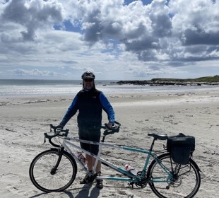 man on beach standing with a tandem bike with a bag on the back of it. The man is wearing a helmet. Its a sunny day. 