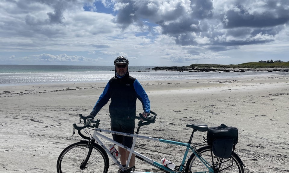 man on beach standing with a tandem bike with a bag on the back of it. The man is wearing a helmet. Its a sunny day. 