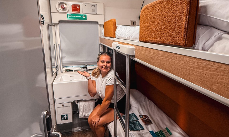 Person seated in a compact train cabin with bunk beds, a sink, and safety instructions on the wall.