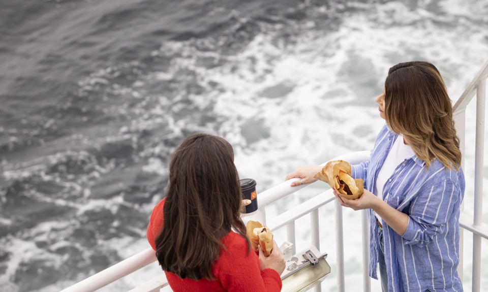 Two females are standing on the deck. One wearing a red sweater and holding a coffee cup, the other wearing a blue and white shirt holding a sandwich