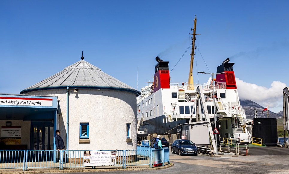 Ferry docked at Port Askaig terminal with vehicles loading, a round white building in front, and mountains under a clear blue sky.