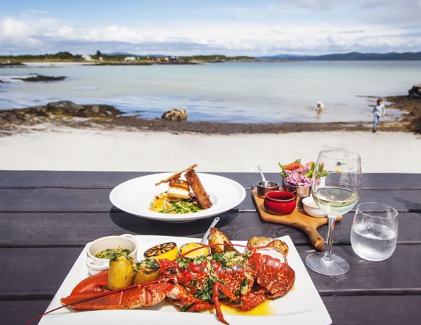 Seafood meal with lobster on a beachside table, clear skies and calm waters in the background.