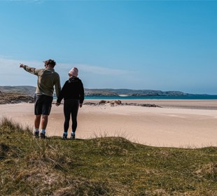Two people holding hands on a grassy hill, looking out at a sandy beach and the ocean beyond.