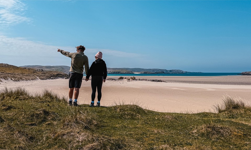 Two people holding hands on a grassy hill, looking out at a sandy beach and the ocean beyond.