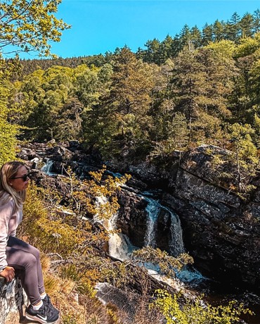 Person in pink hoodie and sunglasses seated on rock ledge, overlooking a forested waterfall under a clear sky.