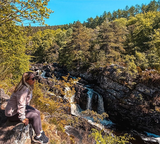 Person in pink hoodie and sunglasses seated on rock ledge, overlooking a forested waterfall under a clear sky.