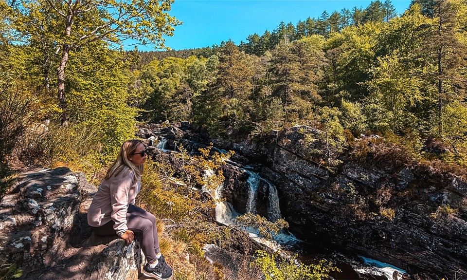 Person in pink hoodie and sunglasses seated on rock ledge, overlooking a forested waterfall under a clear sky.