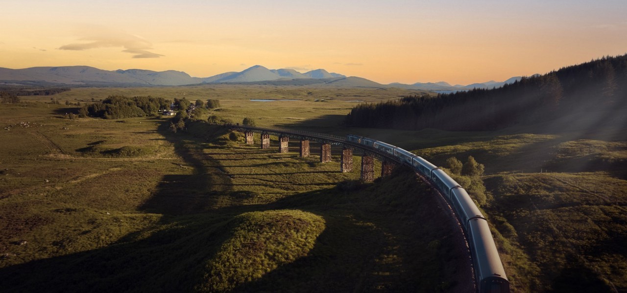 A long passenger train moves across a curved stone viaduct in a green rural landscape at sunset, with mountains in the distance.