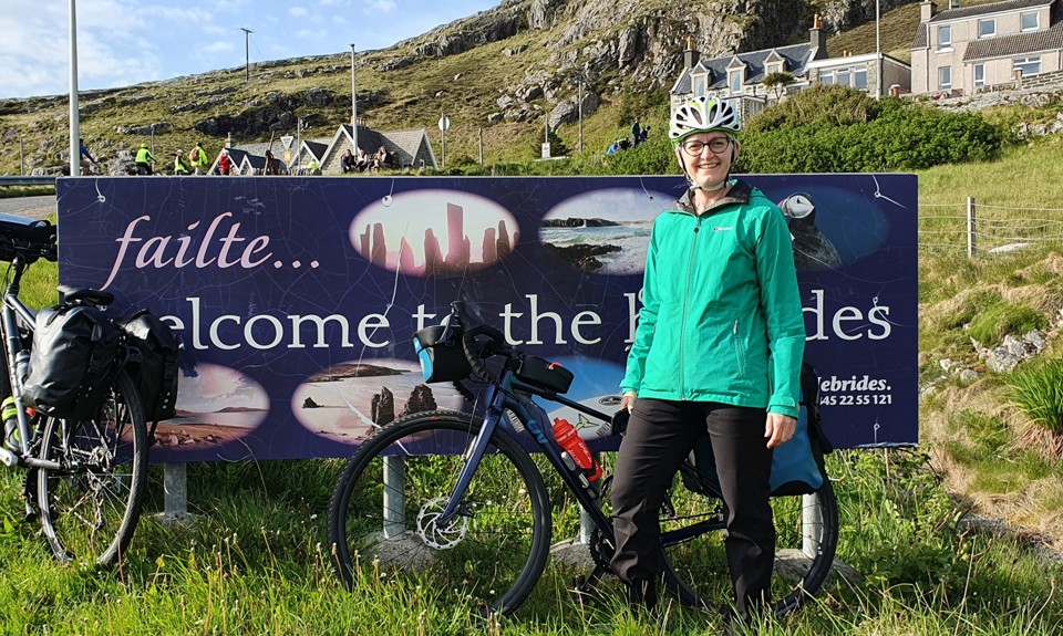 Person in green jacket with bike at 'Welcome to the Hebrides' sign.