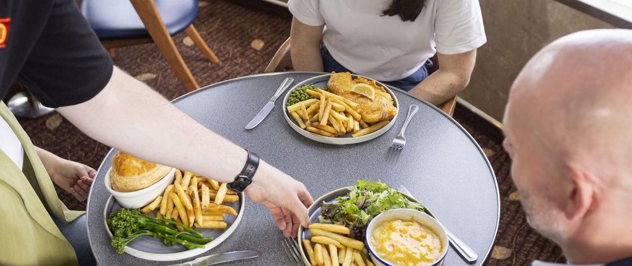 someone placing macaroni cheese, fries and salad in front of a man who's sitting at a table with two females