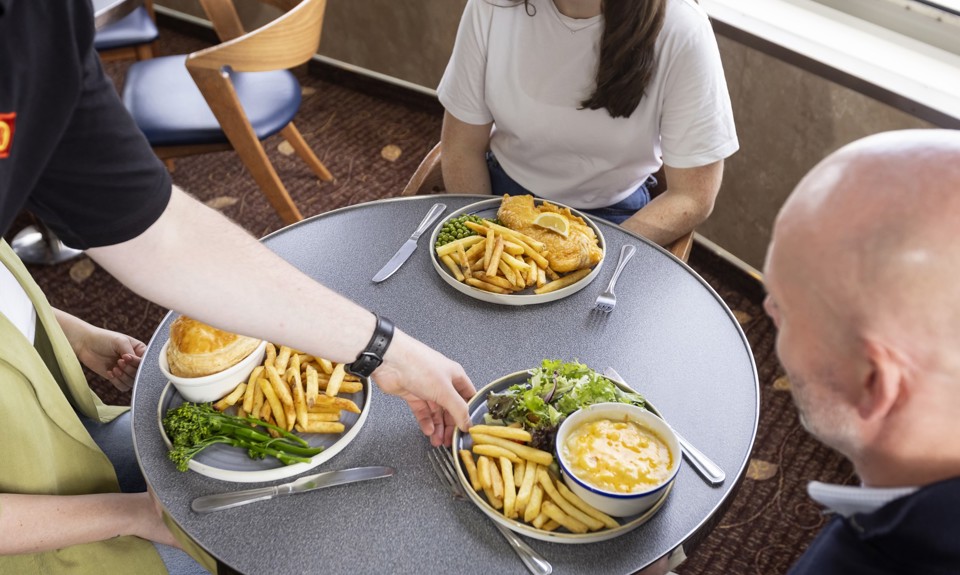 someone placing macaroni cheese, fries and salad in front of a man who's sitting at a table with two females