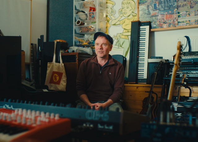 Person seated in a music studio surrounded by keyboards, guitars, recording equipment and artwork.
