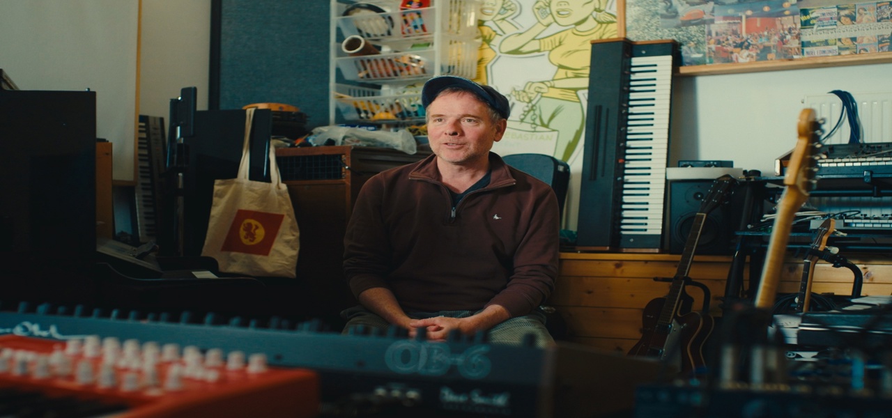 Person seated in a music studio surrounded by keyboards, guitars, recording equipment and artwork.