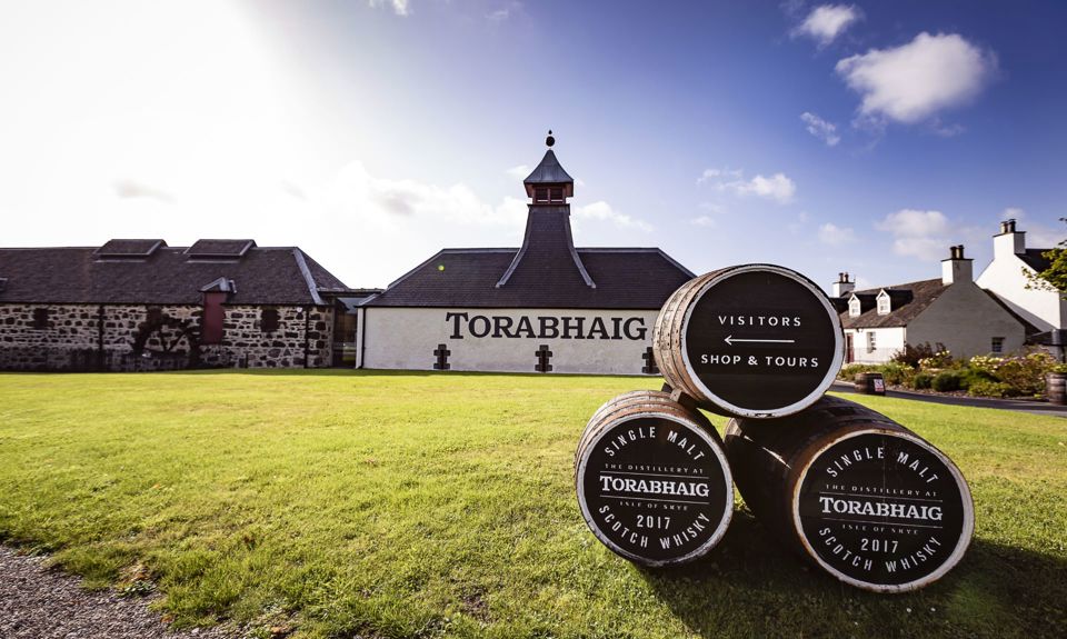 Outside view of Torabhaig Distillery with a white front and dark chimney, with 3 whisky barrels on the grass in the foreground.