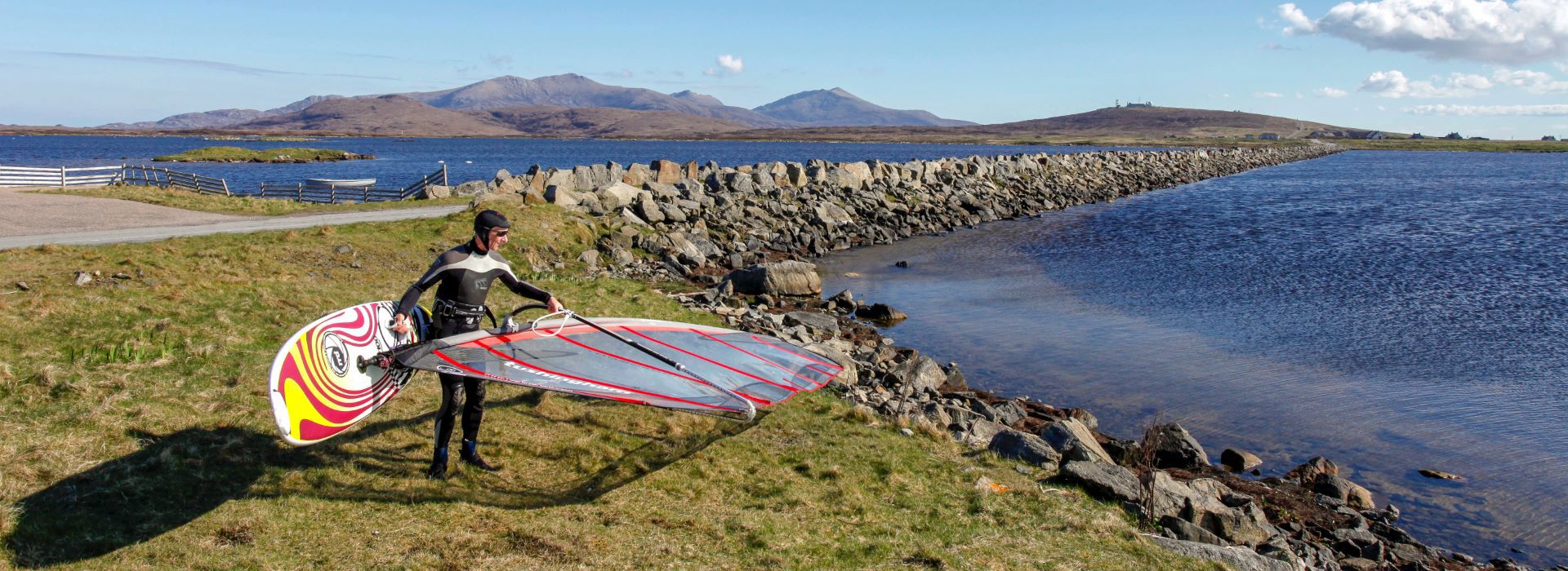 A windsurfer in a wetsuit, carrying their surfboard and sail to Loch Bee, South Uist.