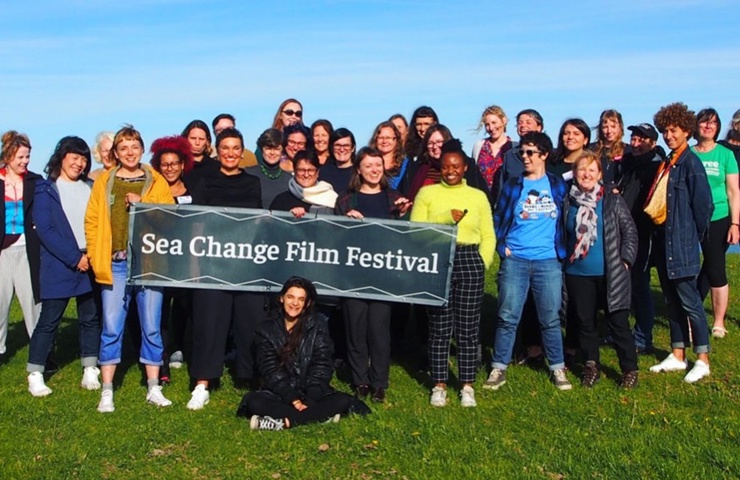 Group of Sea Change Film Festival participants posing outdoors on Tiree, holding a festival banner beside the shoreline.