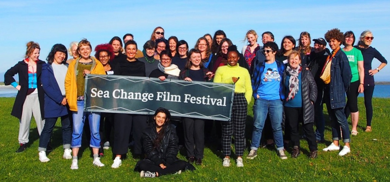 Group of Sea Change Film Festival participants posing outdoors on Tiree, holding a festival banner beside the shoreline.