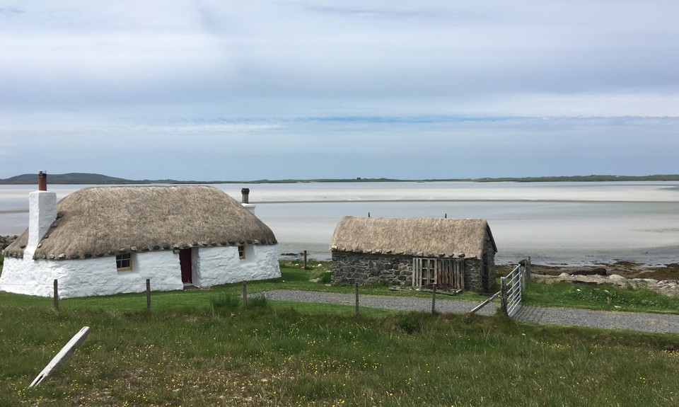 White thatched cottage and stone hut near a calm beach under a cloudy sky.
