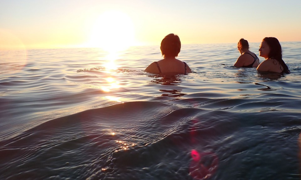 Three people swimming in calm ocean waters during sunset, with the sun low on the horizon casting a warm glow and reflections across the surface.