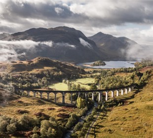The Glenfinnan viaduct spans over the valley with hills and mountains under a cloudy sky.
