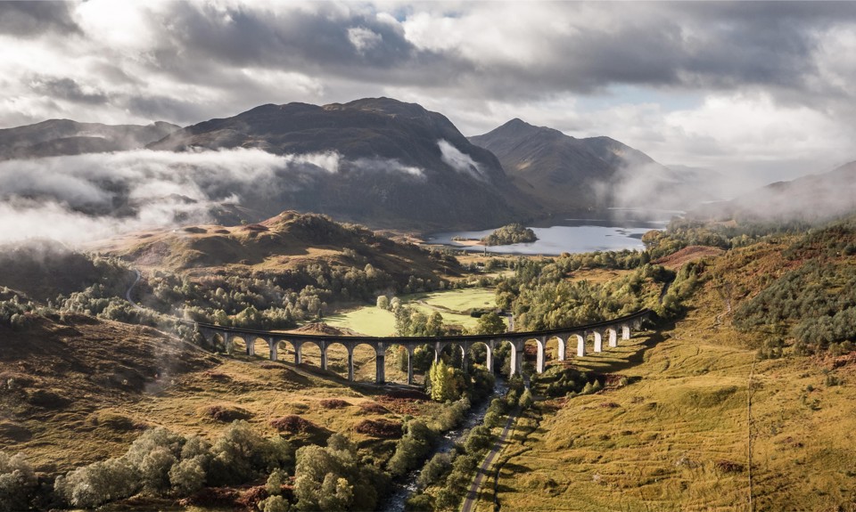 The Glenfinnan viaduct spans over the valley with hills and mountains under a cloudy sky.