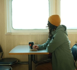 Brunette female with yellow hat sitting at a table looking out a window on a ferry, with a black coffee cup in front of her
