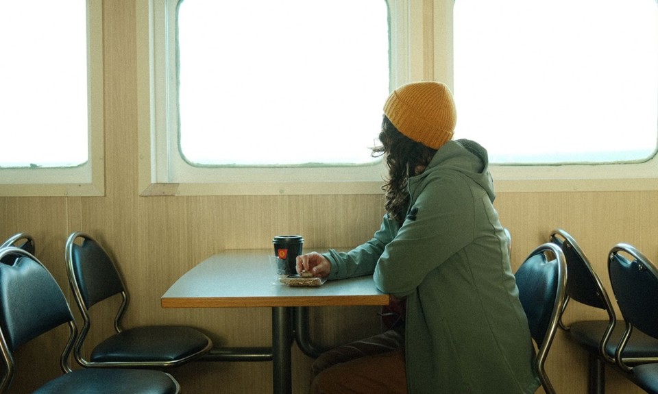 Brunette female with yellow hat sitting at a table looking out a window on a ferry, with a black coffee cup in front of her