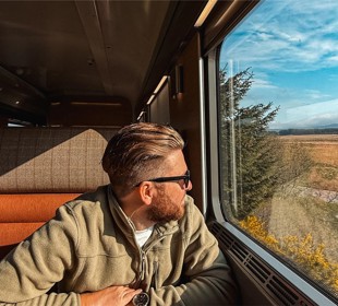 Person in green jacket sits on train, looking out window at scenic landscape with fields, trees, and blue sky.