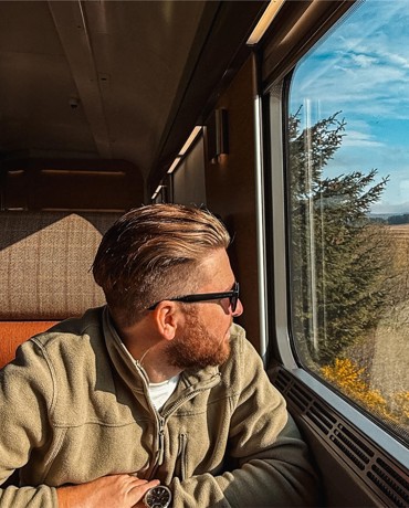Person in green jacket sits on train, looking out window at scenic landscape with fields, trees, and blue sky.
