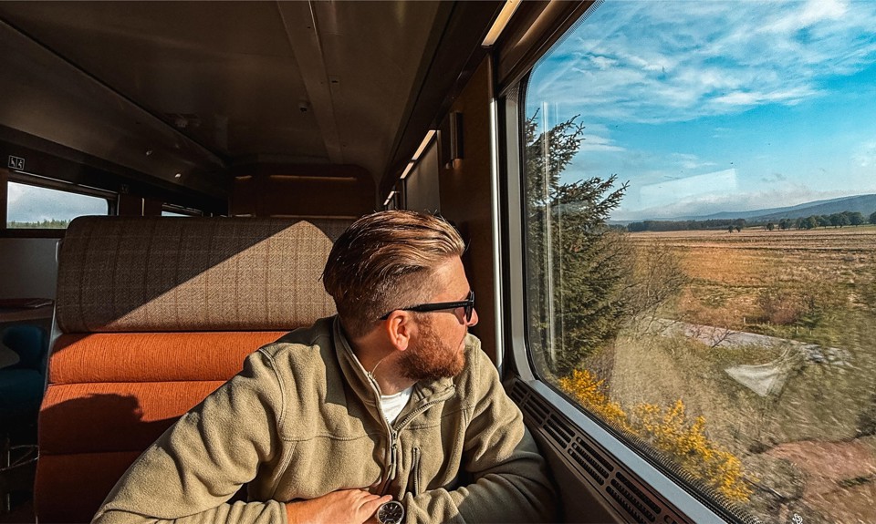 Person in green jacket sits on train, looking out window at scenic landscape with fields, trees, and blue sky.