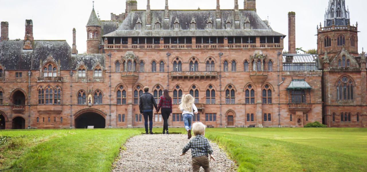 Family walks on a gravel path towards Mount Stuart house, which is a red-brick building with towers, pointed roofs, and many windows.