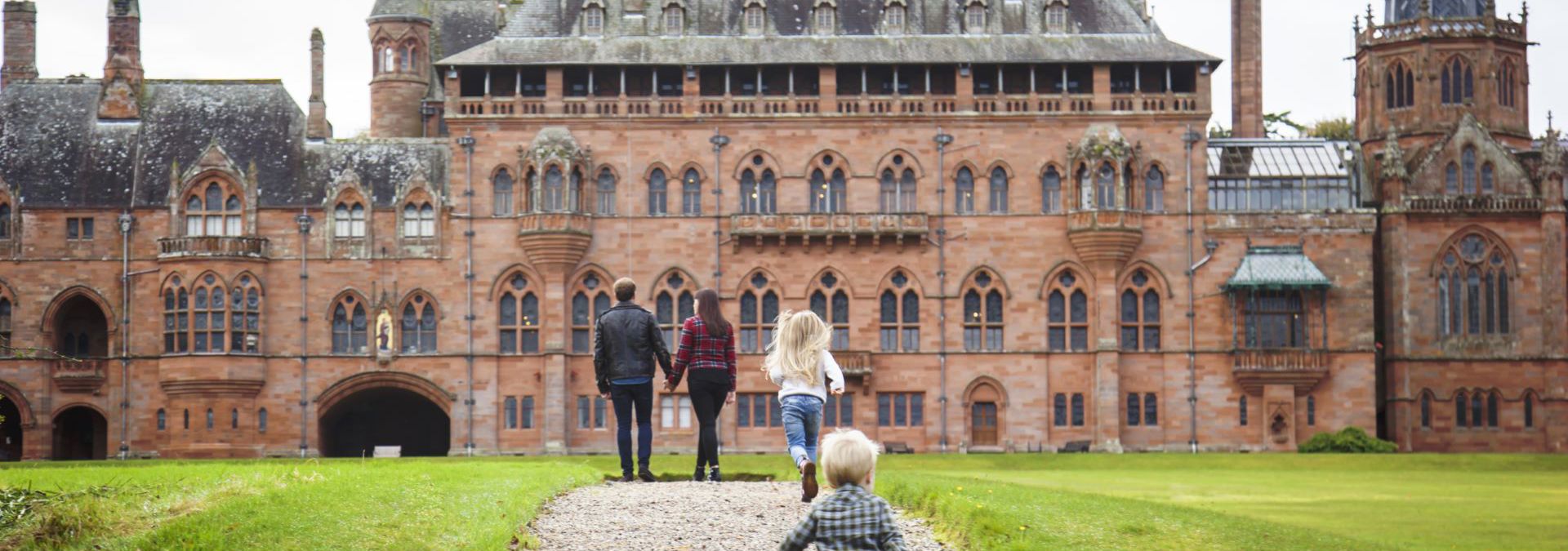 Family walks on a gravel path towards Mount Stuart house, which is a red-brick building with towers, pointed roofs, and many windows.