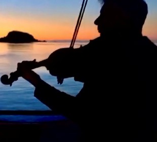 Silhouette of person playing violin on a boat at sunset, with calm water and distant islands.