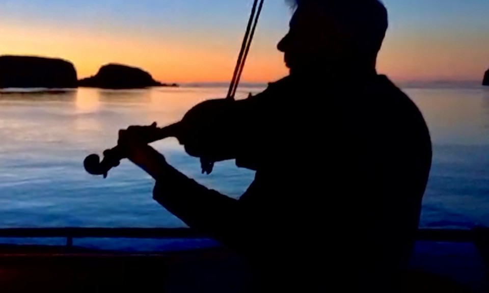 Silhouette of person playing violin on a boat at sunset, with calm water and distant islands.