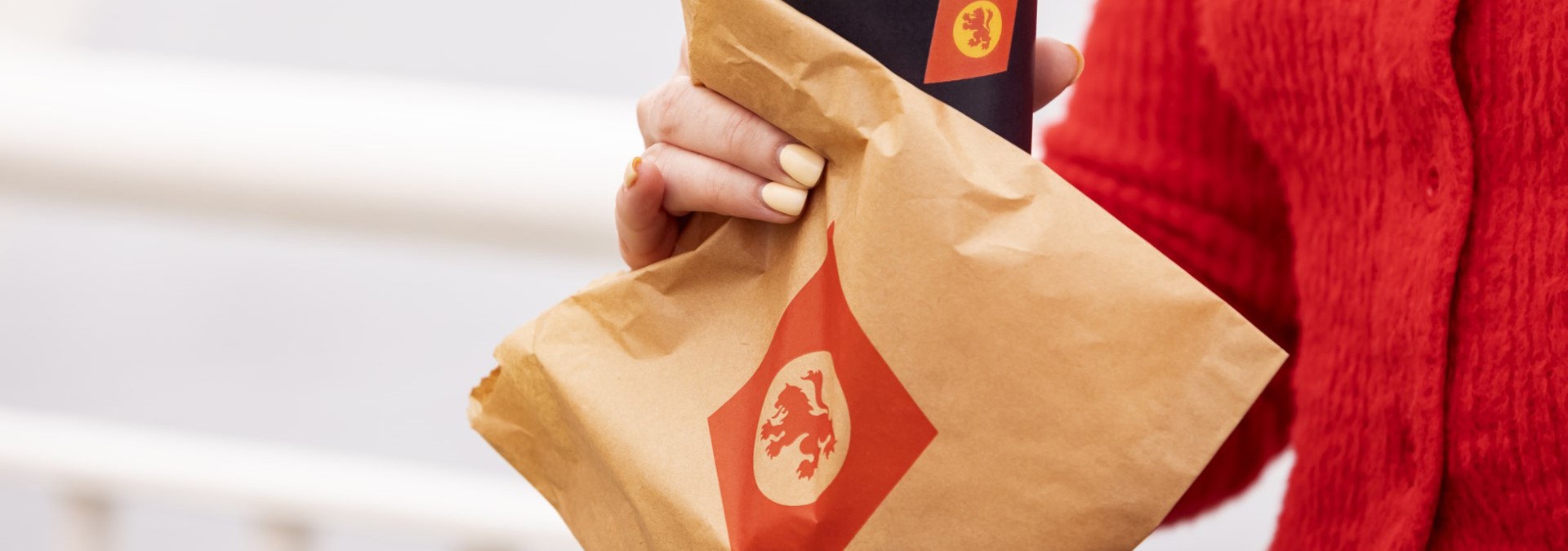 woman in a red sweater holding a brown paper bag with CalMac logo and a cup of coffee in  a black cup