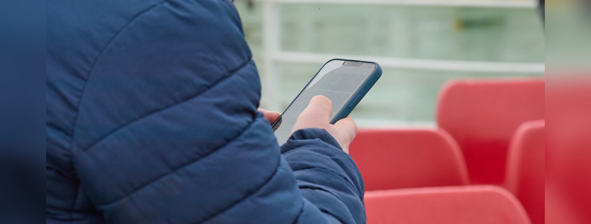 Person using a smartphone while seated on a ferry, with red seating and the vessel deck visible in the background.
